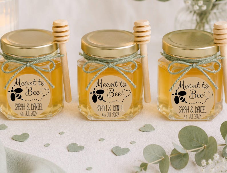 May include: Three hexagonal glass jars filled with golden honey, each topped with a gold lid and a wooden honey dipper. A light green twine bow secures the dipper. A round label reads "Meant to Bee" with a bee graphic and "SARAH & DANIEL 4TH JUL 2027".