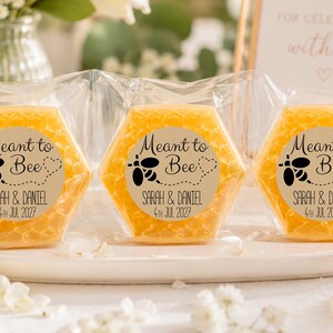 May include: Three hexagonal yellow soaps wrapped in clear plastic, each featuring a circular label with a black bee graphic, the text Meant to Bee, and the names Sarah & Daniel with the date 4th Jul 2027 printed in a clear, dark font.