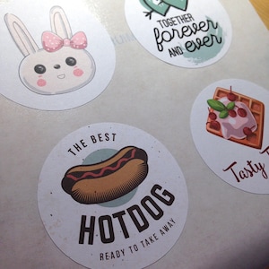 Pu&ograve; includere: Un foglio di adesivi circolari con vari disegni: un coniglio bianco con un fiocco rosa, due cuori verdi con una freccia e il testo "TOGETHER forever AND ever", un waffle con ciliegie e un hot dog con il testo "THE BEST HOTDOG READY TO TAKE AWAY".