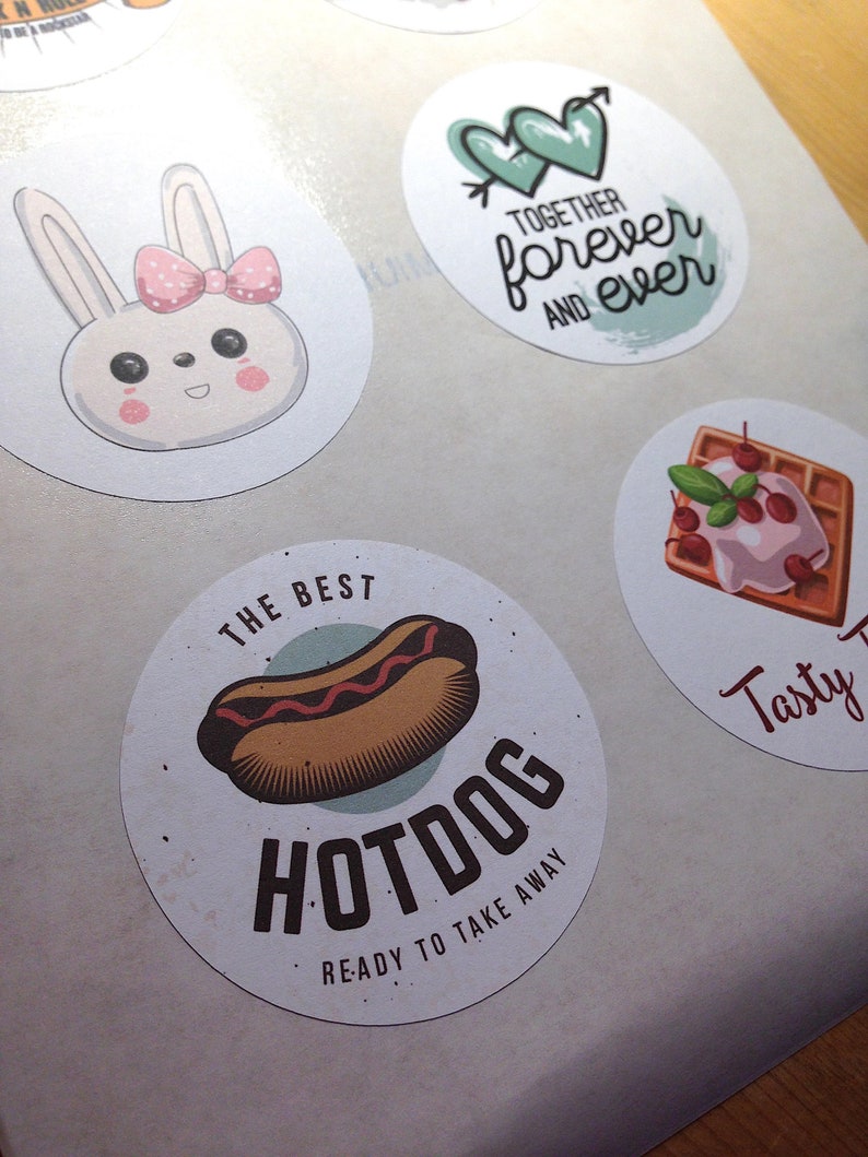 Pu&ograve; includere: Un foglio di adesivi circolari con vari disegni: un coniglio bianco con un fiocco rosa, due cuori verdi con una freccia e il testo "TOGETHER forever AND ever", un waffle con ciliegie e un hot dog con il testo "THE BEST HOTDOG READY TO TAKE AWAY".