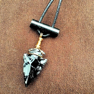 May include: A black obsidian arrowhead pendant on a black leather cord with a wooden toggle clasp. The arrowhead is wrapped in gold wire.