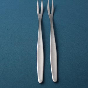 May include: Two silver-toned, pronged forks with long, slender handles. The forks are positioned vertically on a teal background.