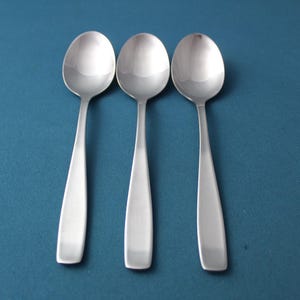 May include: Three stainless steel spoons with oval bowls and elongated, flat handles. The spoons are arranged vertically against a teal background, reflecting light. The spoons are likely for eating.
