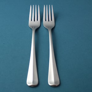 May include: Two silver forks with a simple design are placed side by side on a blue surface.
