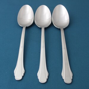 May include: Three stainless steel spoons arranged on a blue surface. Each spoon has a rounded bowl and a long, decorative handle with a flared, fan-like design at the base. The spoons are evenly spaced and reflect the light.