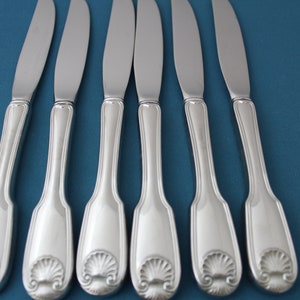 May include: Six silver dinner knives with ornate handles featuring a shell design. The knives are arranged in a row on a blue background.
