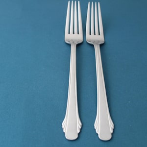 May include: Two stainless steel forks with a decorative handle design. The forks are positioned vertically against a teal background. Each fork has four tines and a long, slender handle.