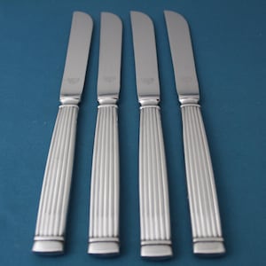 May include: Four silver butter knives with a ridged handle design. The blades are smooth and curved. The knives are arranged diagonally against a blue background, suggesting a formal dining setting.