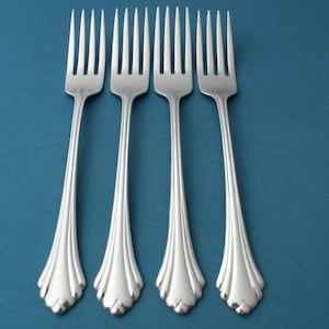 May include: Four silver forks with a decorative handle design are arranged on a teal background. The forks have tines and a detailed, ornate handle. The image is a close-up shot of the silverware.