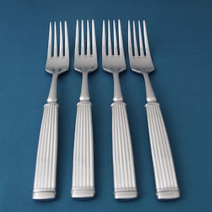 May include: Four silver forks with a classic design, arranged on a blue background. Each fork has a tined head and a handle with vertical grooves. The forks are evenly spaced and angled slightly.