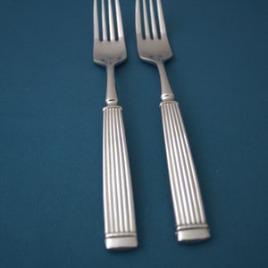 May include: Two stainless steel forks with a fluted handle design. The forks are set against a teal background, creating a contrast that highlights the silverware's polished finish. Ideal for formal dining.