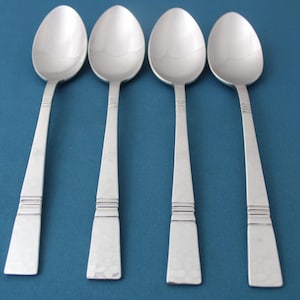 May include: Four silver spoons with oval bowls and textured handles are arranged against a blue background. The spoons have a simple, elegant design, suitable for everyday use or special occasions. The handles have a hammered texture.