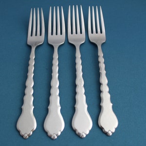May include: Four silver forks with ornate handles are arranged on a blue surface. Each fork has four tines and a decorative handle with a teardrop-shaped end. The forks are evenly spaced and reflect the light.