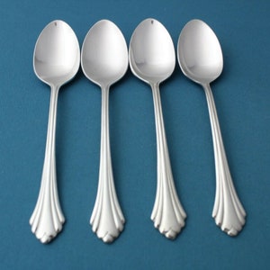 May include: Four silver spoons arranged on a blue background. Each spoon has an oval bowl and a decorative handle with a stylized floral design. The spoons are made of a shiny, reflective metal.