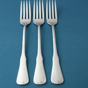 May include: Three silver forks with a classic design are arranged on a teal background. Each fork has a long, slender handle that widens at the base and a four-tine head. The forks are evenly spaced.