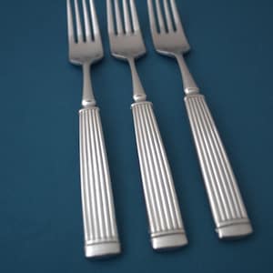May include: Three silver forks with a fluted handle design are arranged diagonally on a blue surface. The forks have a classic, elegant appearance, suitable for formal dining. The tines are visible, and the handles have a detailed, textured pattern.