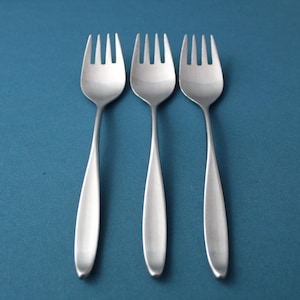 May include: Three stainless steel forks with a simple, modern design. The forks are arranged vertically against a solid teal background. Each fork has a long, slender handle and four tines.