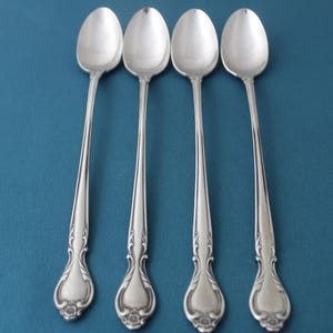 May include: Four silver spoons with long handles and ornate floral designs on the handles and bowls. The spoons are arranged vertically against a teal background, showcasing their polished, reflective surfaces. The spoons are likely for serving or eating.