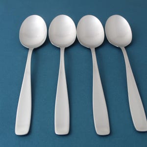 May include: Four stainless steel spoons with oval bowls and elongated, flat handles are arranged on a teal background. The spoons are reflective, and the handles have a slight taper. The spoons are for serving food.