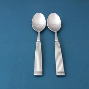 May include: Two silver spoons with oval bowls and vertically ribbed handles are displayed against a blue background. The spoons have a classic design, suitable for serving or eating. The spoons are made of a shiny, reflective metal.