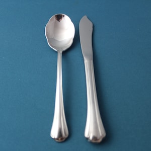 May include: A silver spoon and butter knife set against a teal background. The spoon has a rounded bowl with a scalloped edge, and the knife has a curved blade. Both utensils have fluted handles.