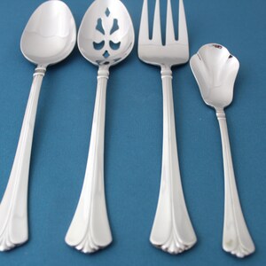 May include: Set of four silver-toned flatware pieces, including a serving spoon, a slotted serving spoon, a fork, and a butter knife. The pieces have ornate handles with a decorative design.