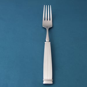 May include: A silver fork with four tines and a long, ridged handle. The fork is centered against a solid teal background, creating a simple and elegant presentation. The handle has a rectangular base.