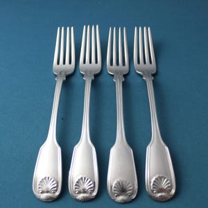 May include: Four silver forks with a shell design on the handle, arranged on a teal background. The forks have three tines and a long, slender handle. The shell design adds a decorative element to the silverware.