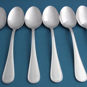 May include: Six silver spoons with rounded bowls and a simple design. The spoons are arranged in a row on a blue background.