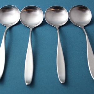 May include: Four stainless steel soup spoons with rounded bowls and long, slender handles. The spoons are arranged in a row against a teal background, reflecting light. The silverware is designed for serving soup or other liquid dishes.