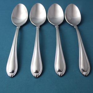 May include: Four stainless steel spoons arranged on a teal background. Each spoon has a teardrop-shaped bowl and a long, slender handle with a decorative ridge. The spoons are evenly spaced, reflecting light.