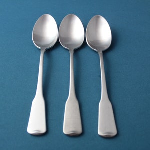 May include: Three silver spoons with oval bowls and rectangular handles are arranged on a teal background. The spoons are made of a shiny, reflective metal, and the handles have a slightly flared shape.