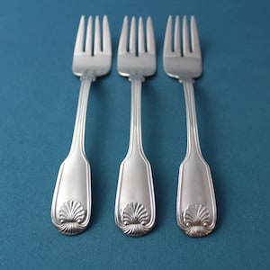 May include: Three silver forks with ornate handles featuring a shell design. The forks are arranged in a row on a blue background.