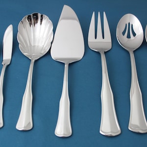 May include: Set of seven silver-toned serving utensils, including a butter knife, a serving spoon, a cake server, a slotted spoon, a serving fork, and two spoons with scalloped edges.