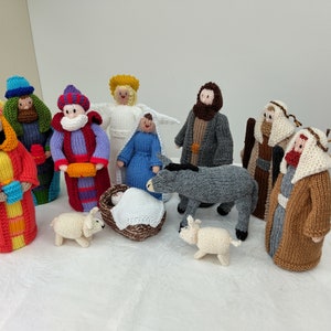 May include: A knitted nativity set with figures including the Three Wise Men, Mary, Joseph, baby Jesus, an angel, a donkey, and sheep. The figures are brightly coloured with details such as crowns, robes, and beards. The scene is set on a white surface.