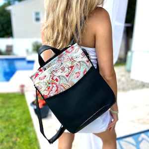 Puede incluir: Una mochila negra con una solapa de estampado paisley. La solapa está doblada sobre la parte superior de la bolsa y asegurada con una cremallera. La bolsa tiene correas ajustables.