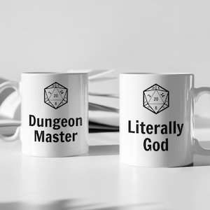 May include: Two white ceramic mugs with black text and a black and white image of a twenty-sided die. The mug on the left says "Dungeon Master" and the mug on the right says "Literally God".