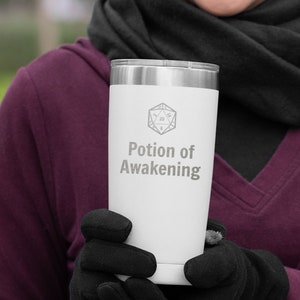 May include: A white stainless steel travel mug with a black lid and a gray graphic of a twenty-sided die and the text "Potion of Awakening".