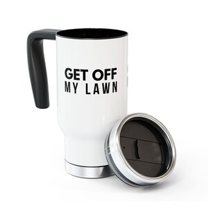 May include: A white travel mug with a black handle and a black interior. The text GET OFF MY LAWN is printed in black on the front. A matching black lid with a sliding closure is positioned next to the base of the mug on the right side.
