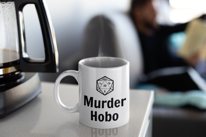 K&ouml;nnte beinhalten: Wei&szlig;e Keramik-Kaffeetasse mit einer schwarz-wei&szlig;en Illustration eines zwanzigseitigen W&uuml;rfels und dem Text "Murder Hobo".