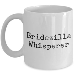 May include: White ceramic coffee mug with black text that reads "Bridezilla Whisperer".