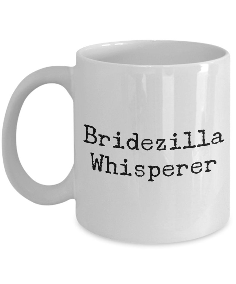 May include: White ceramic coffee mug with black text that reads "Bridezilla Whisperer".