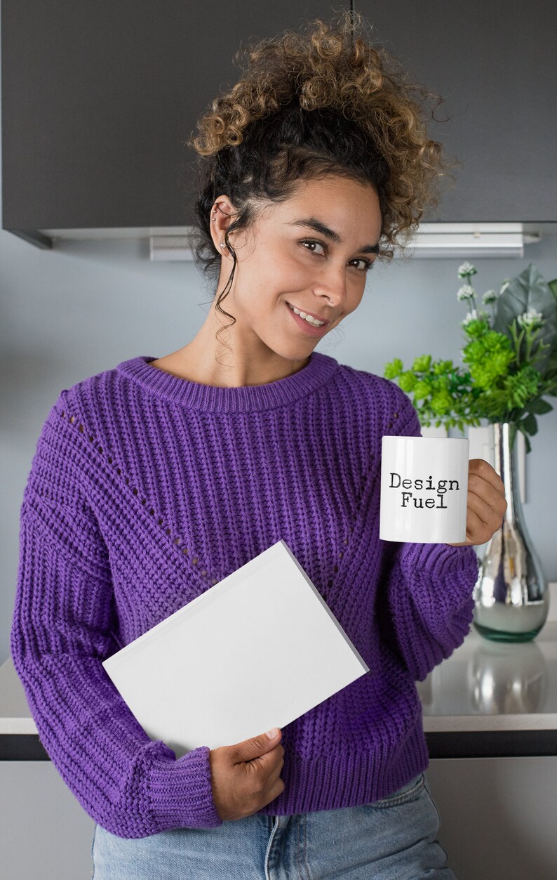 May include: A woman wearing a purple sweater holds a white mug with the text "Design Fuel" and a white book.