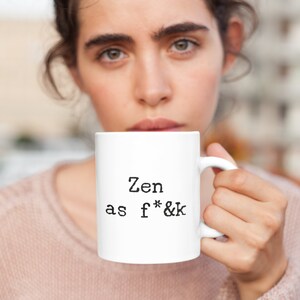May include: White ceramic mug with the text "Zen as f*&k" in black. The mug is held by a person wearing a pink sweater. The mug has a standard handle and a cylindrical shape. The text is centered on the mug.