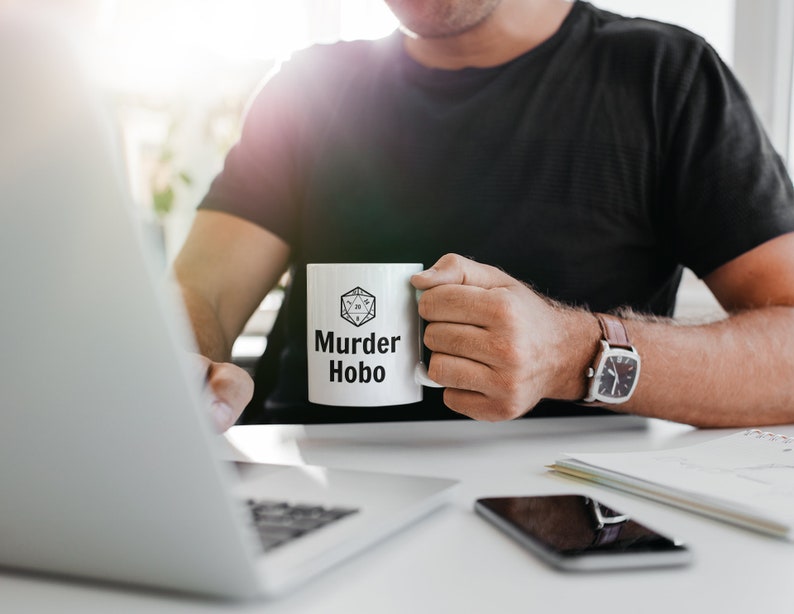 K&ouml;nnte beinhalten: Eine wei&szlig;e Keramiktasse mit einer schwarz-wei&szlig;en Grafik eines W&uuml;rfels und dem Text "Murder Hobo".
