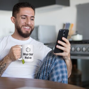 K&ouml;nnte beinhalten: Eine wei&szlig;e Keramiktasse mit einer schwarz-wei&szlig;en Grafik eines W&uuml;rfels mit dem Text "Murder Hobo" darauf. Eine Person h&auml;lt die Tasse in ihrer rechten Hand und schaut auf ein schwarzes Smartphone in ihrer linken Hand.