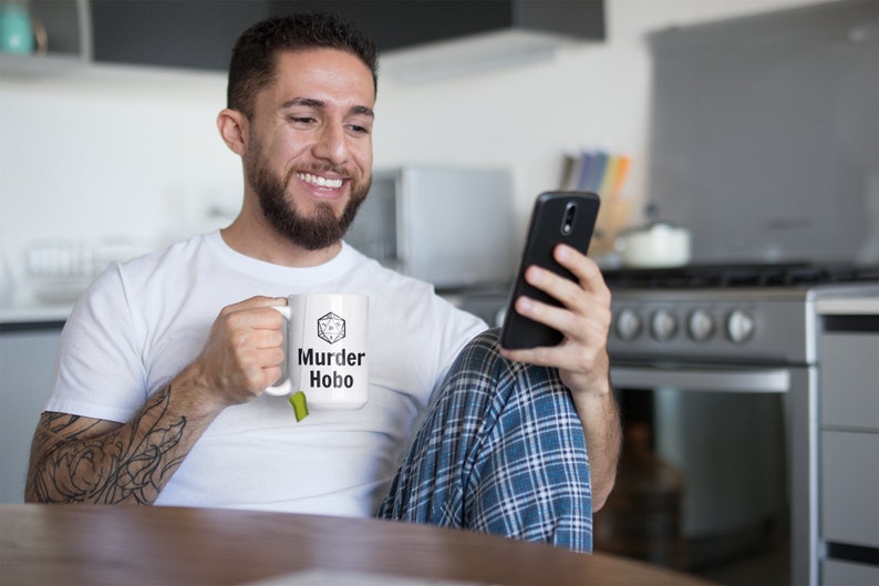 K&ouml;nnte beinhalten: Eine wei&szlig;e Keramiktasse mit einer schwarz-wei&szlig;en Grafik eines W&uuml;rfels mit dem Text "Murder Hobo" darauf. Eine Person h&auml;lt die Tasse in ihrer rechten Hand und schaut auf ein schwarzes Smartphone in ihrer linken Hand.