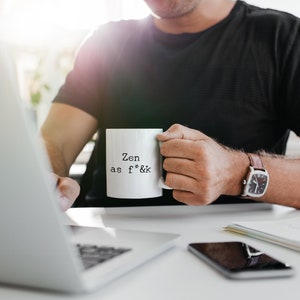 May include: A white ceramic mug with the text "Zen as f*&k" in black, held by a person. The person is wearing a black shirt and a watch with a brown leather strap. A laptop and smartphone are on the desk.