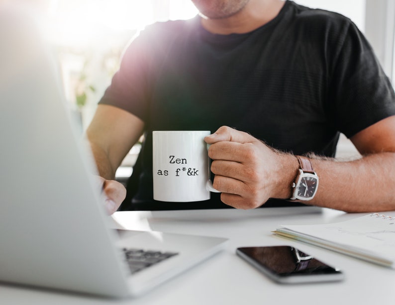May include: A white ceramic mug with the text "Zen as f*&k" in black, held by a person. The person is wearing a black shirt and a watch with a brown leather strap. A laptop and smartphone are on the desk.