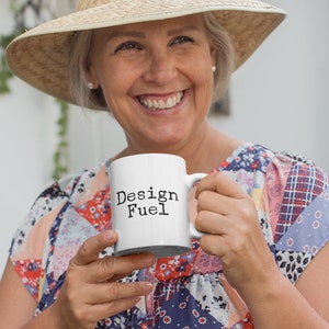 May include: A white ceramic mug with the text "Design Fuel" in black lettering. A person is holding the mug and smiling.